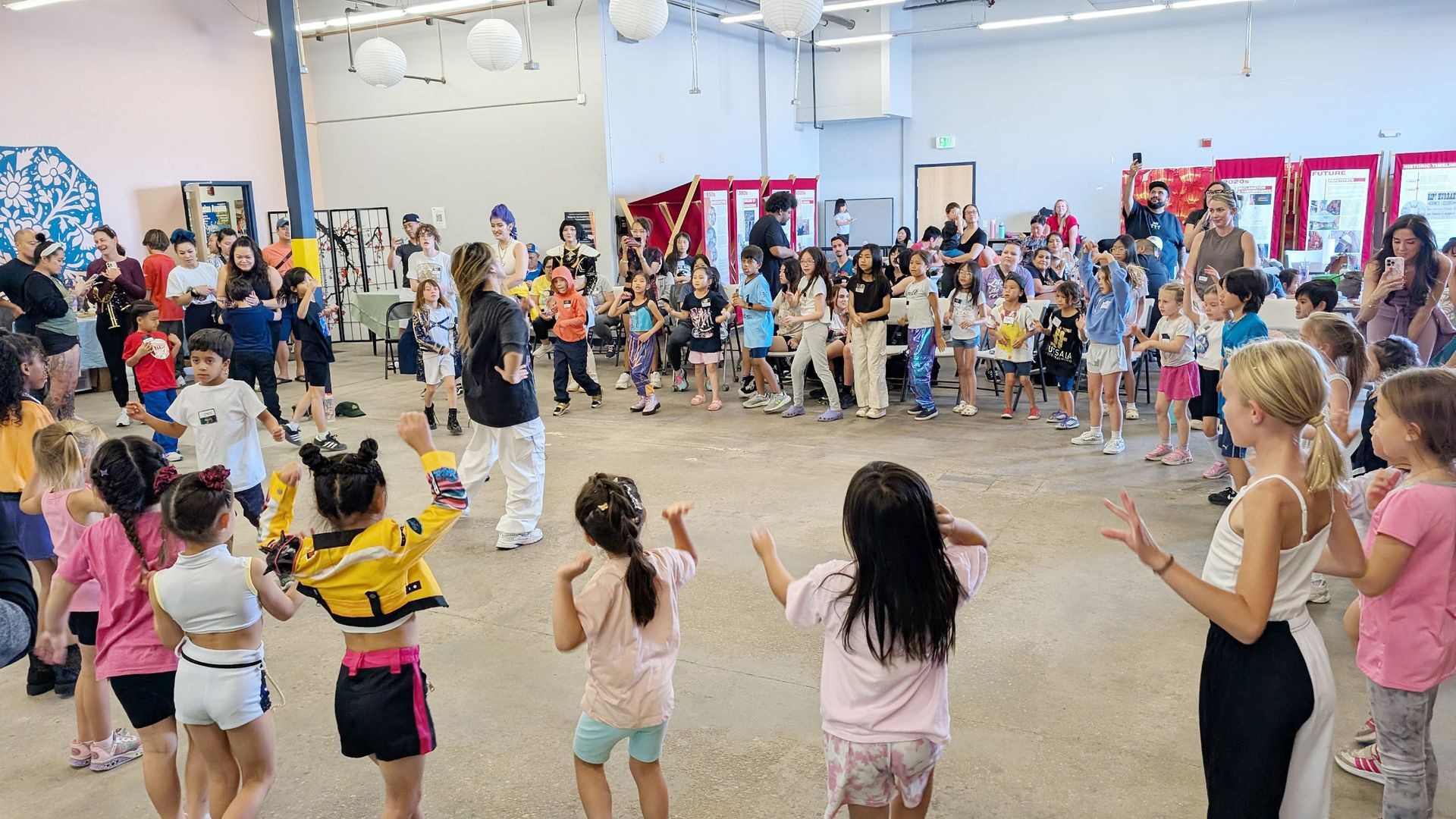 Children in a large room, some dancing. Others watch, with adults in the background.
