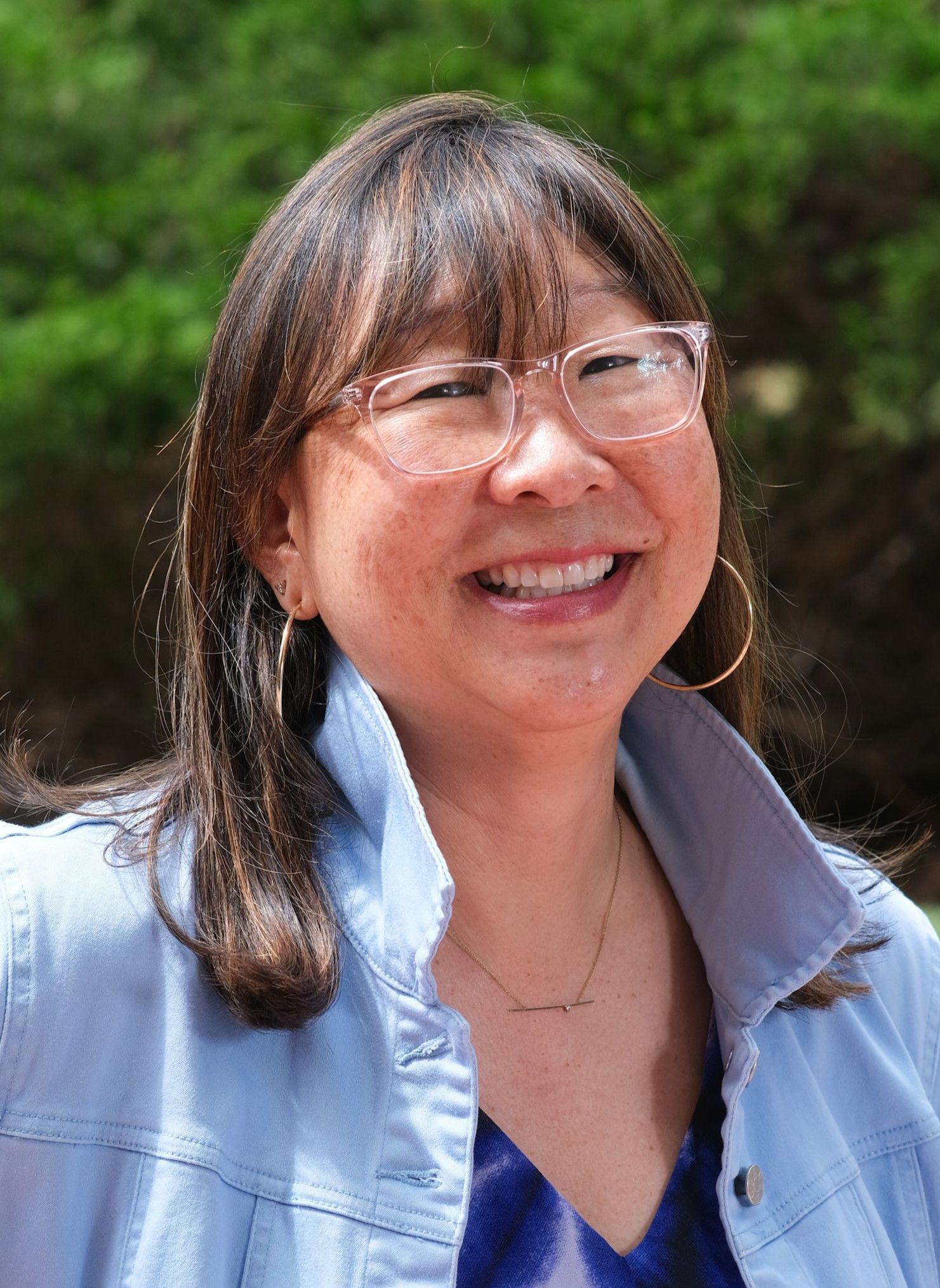 Stacey Shigaya smiling, wearing glasses, denim jacket, and hoop earrings, outdoors.