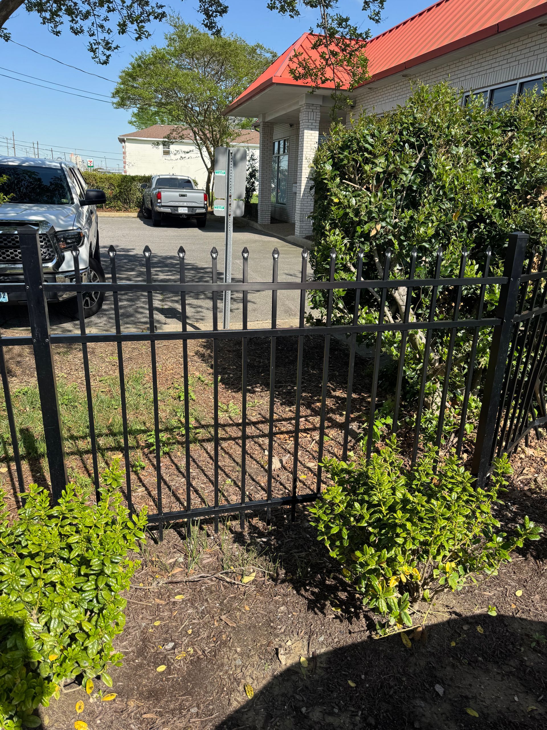 Black metal fence with bushes in front, cars and building in background. Sunny day.
