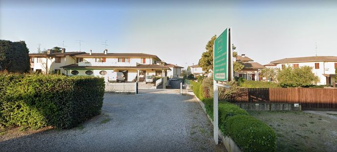 Un vialetto di ghiaia conduce a un edificio a più piani di colore chiaro, con un'insegna verde e siepi ben curate in primo piano.