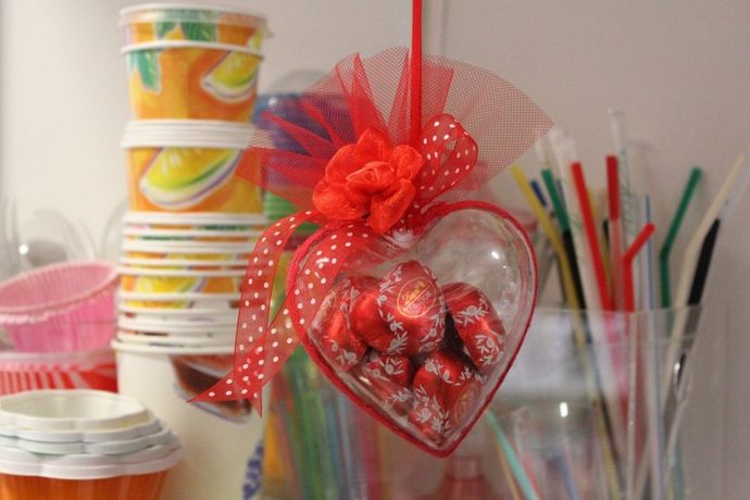 Un contenitore trasparente a forma di cuore, riempito di caramelle incartate in rosso, decorato con un nastro rosso e una rosa di stoffa.