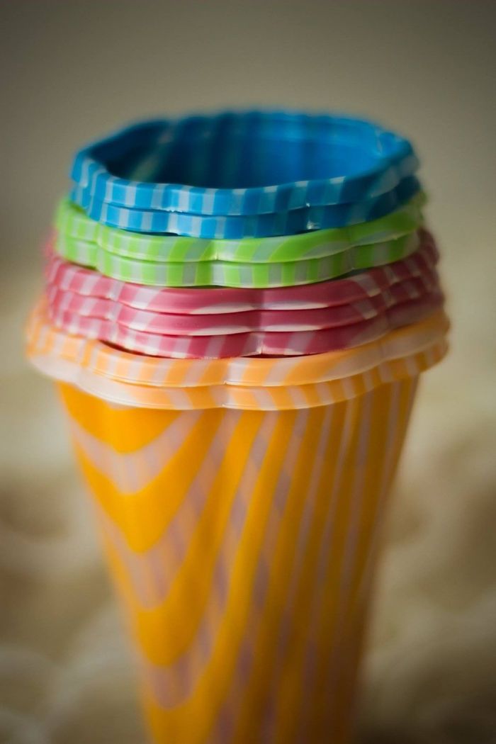 Una pila di quattro bicchieri di plastica colorati con bordi smerlati, nei colori blu, verde, rosa e arancione, su uno sfondo sfocato.