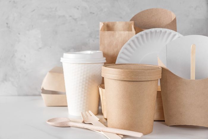 Su un tavolo bianco sono disposti diversi contenitori per alimenti ecocompatibili e biodegradabili, piatti di carta e posate di legno.