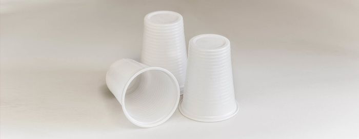 Tre bicchieri di plastica bianca usa e getta, uno appoggiato su un fianco e due in posizione verticale, su uno sfondo chiaro e uniforme.