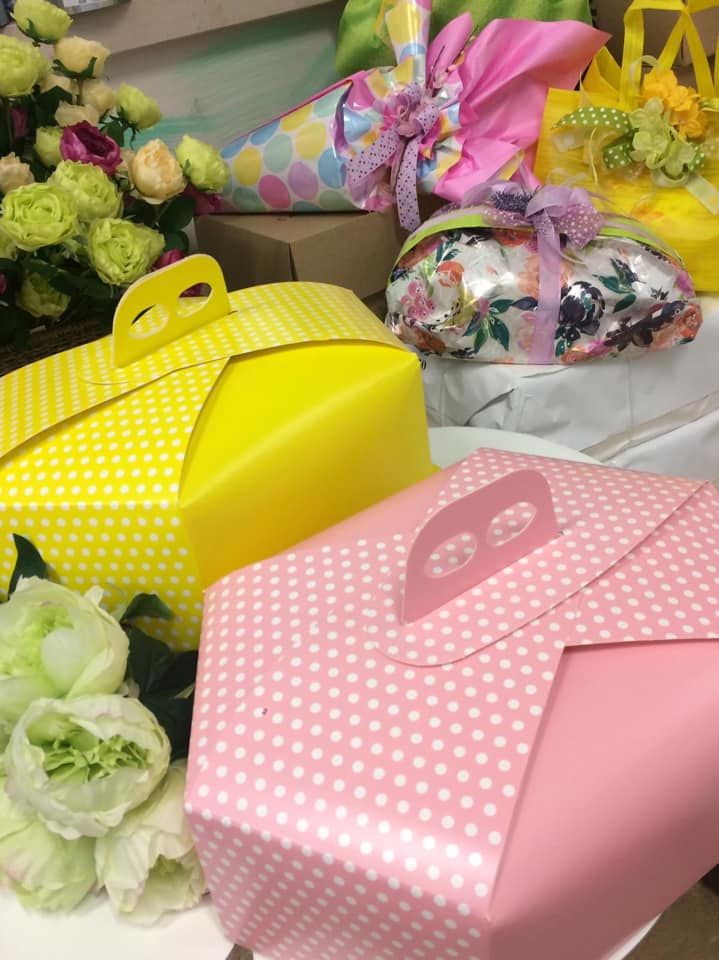 Scatole regalo dai colori pastello con manici gialli e rosa sono disposte accanto a composizioni floreali e pacchi regalo.