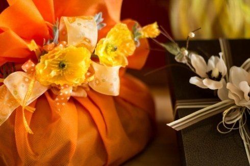 Un regalo arancione avvolto in tessuto e decorato con fiori gialli, accanto a un regalo scuro con fiori bianchi e nastro.