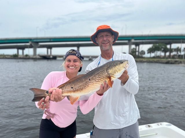 Two people hold a large redfish on a boat near a bridge.