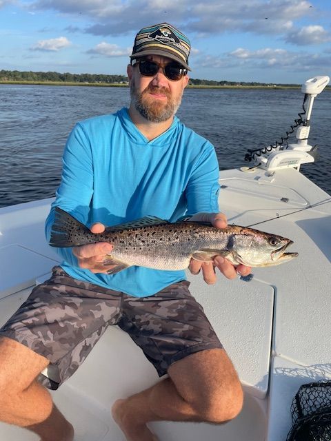 Man on boat holding spotted fish. He wears sunglasses, blue shirt, shorts. Water and sky background.