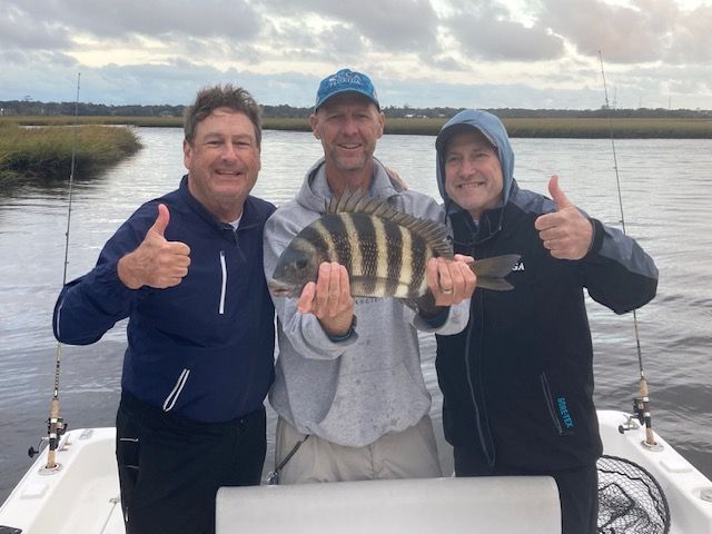 Three men on a boat, holding a striped fish. Two give thumbs up. Overcast sky, marsh in background.