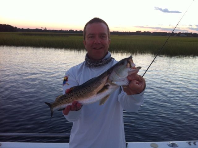 Man holding a redfish he caught, smiling, near water at dusk.