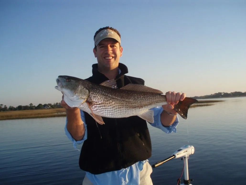 Man on boat smiles, holding a large redfish. Blue water and sky.