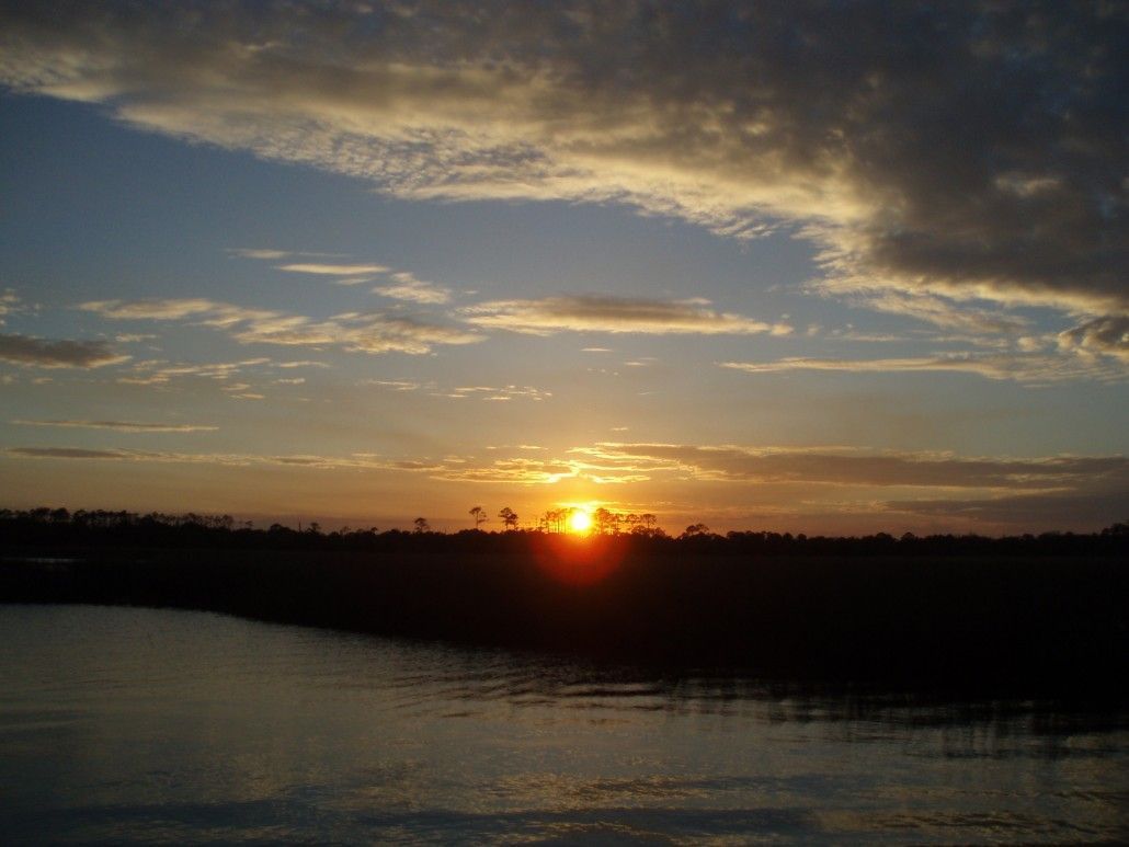 Sunset over dark treeline and water, with orange and yellow sun behind trees, blue and gray sky.