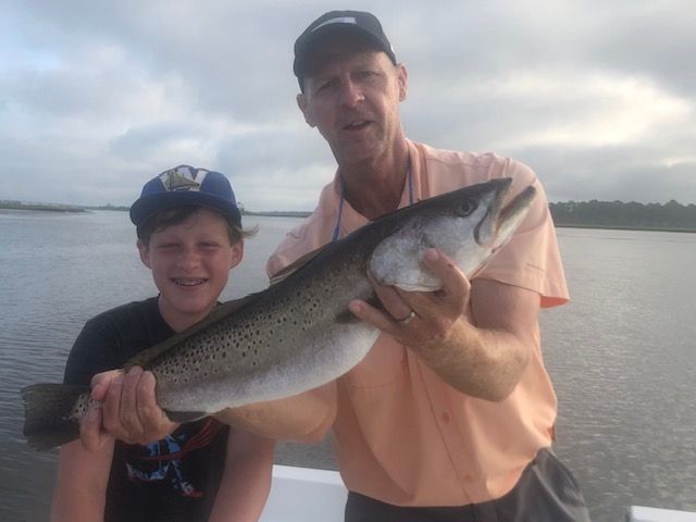 Man and child holding a large speckled trout on a boat, against a water and sky backdrop.
