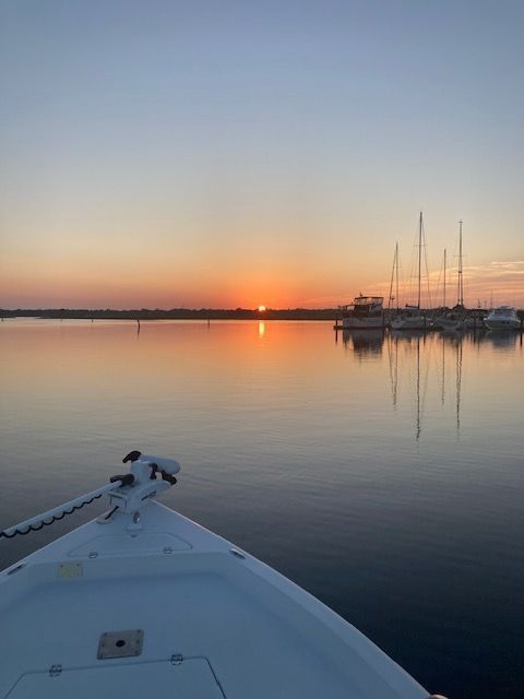 Boat on calm water, sunrise over a marina with docked sailboats.