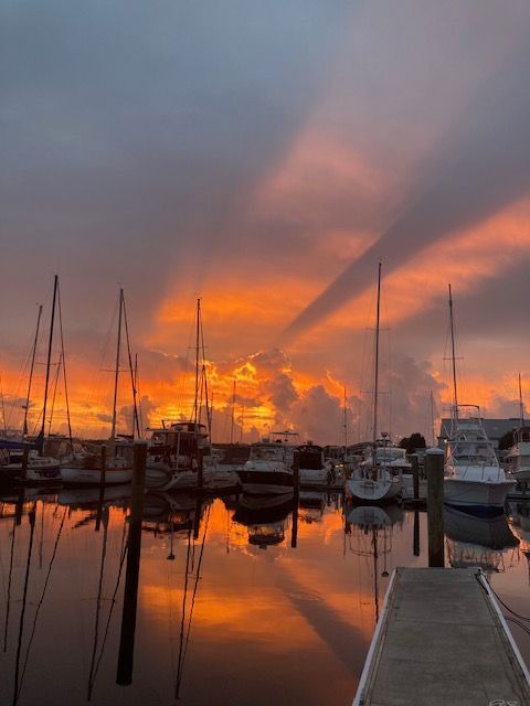 Orange sunset over a marina, boats and their reflections in water, with rays of light through clouds.