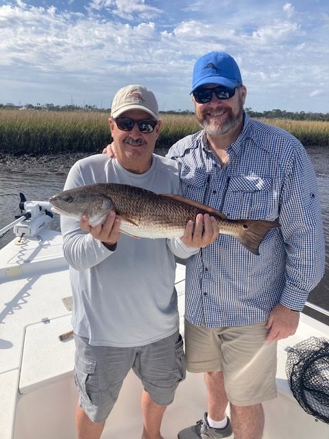 Two men on a boat hold a large redfish. Salt marsh and cloudy sky in background.
