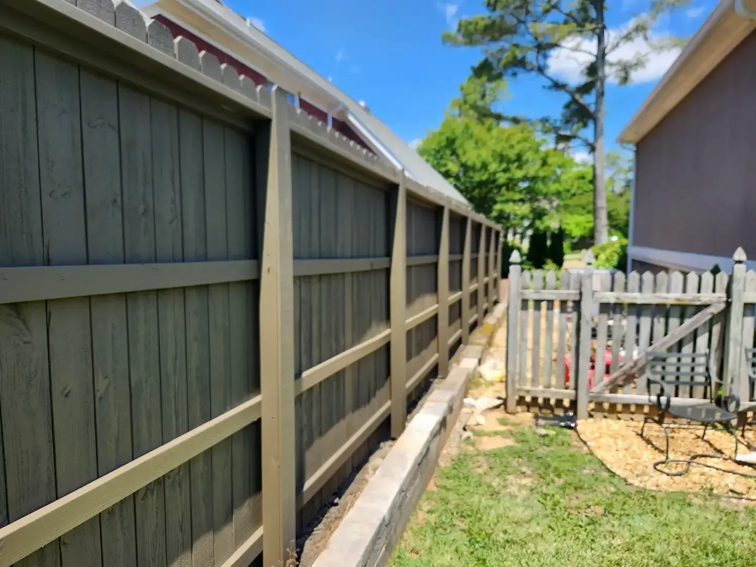 Long wooden fence along a narrow side yard beside a house, with a small white gate and sunny sky.