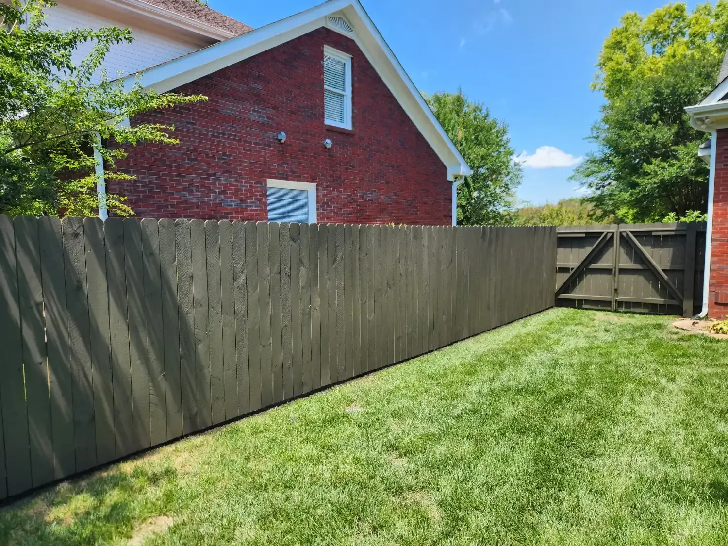 Backyard with green lawn, wooden fence, and red house under a blue sky