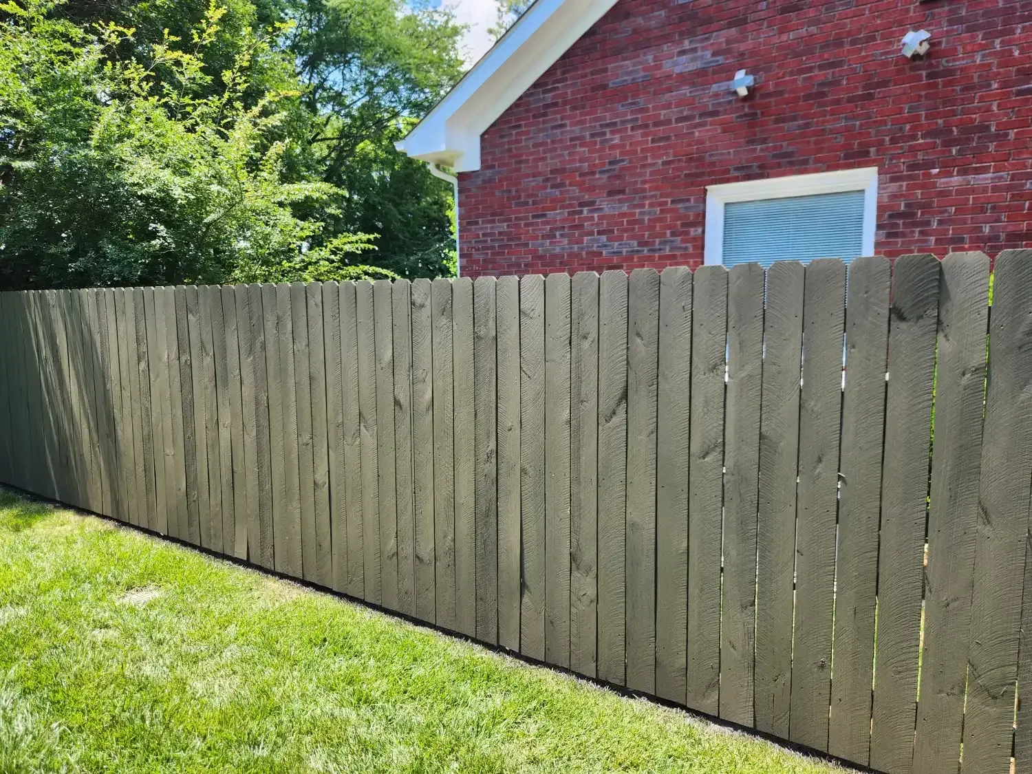 Weathered wooden fence beside a green lawn with a red brick house and trees behind it