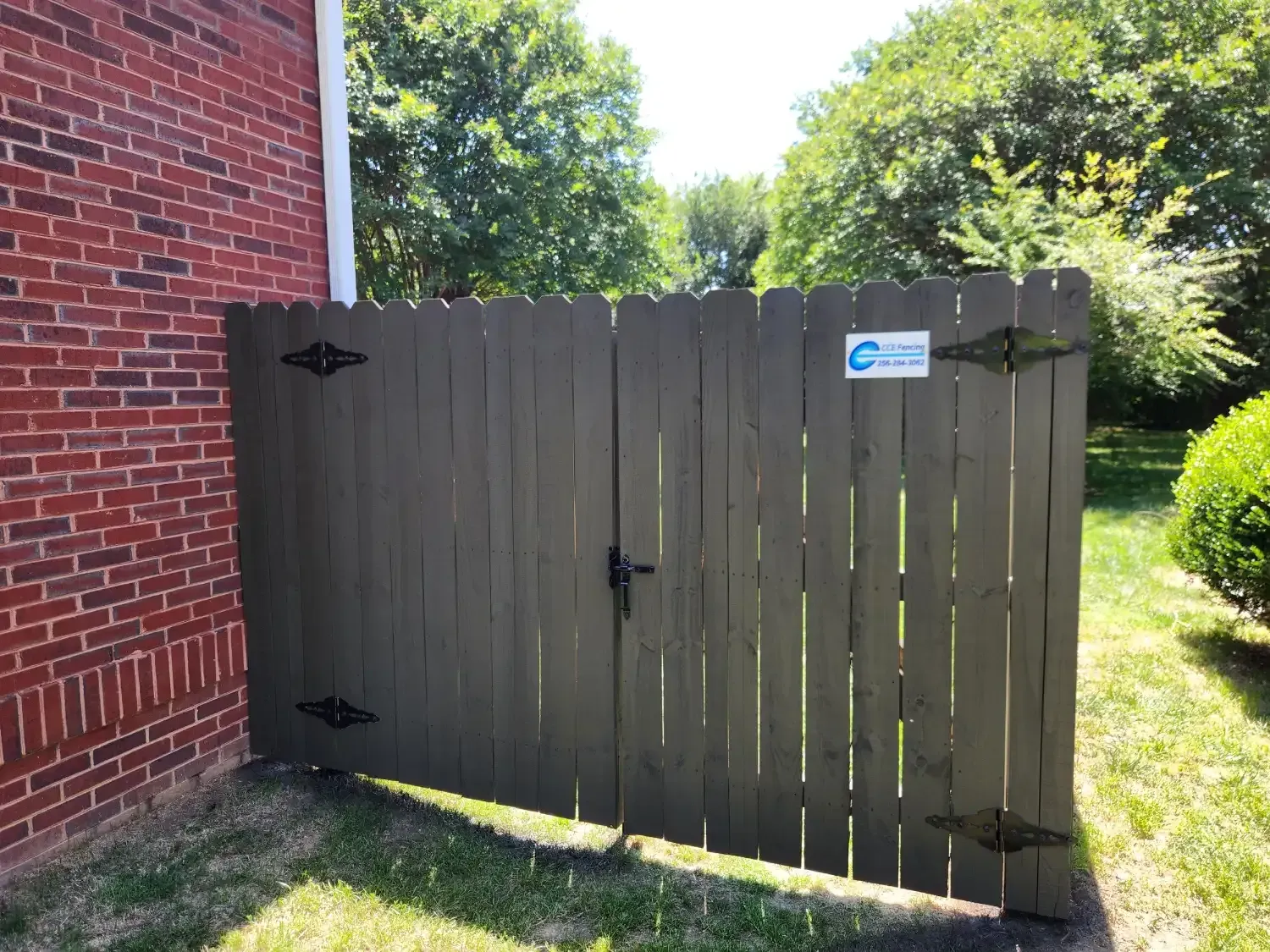 Brown wooden gate beside a red brick building, opening to a grassy yard with trees.
