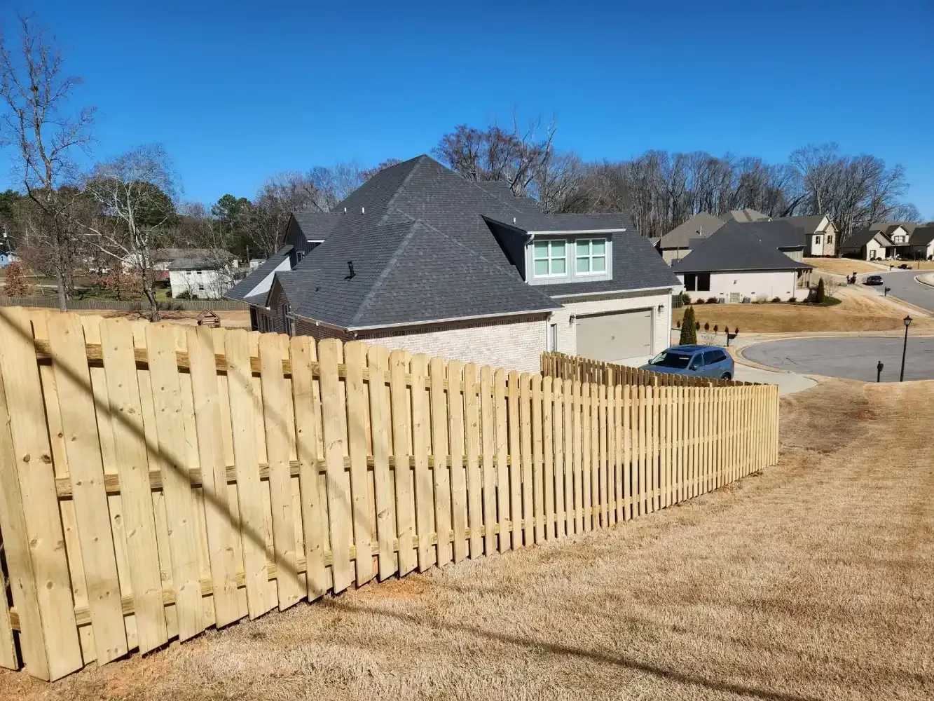 Wooden privacy fence along a grassy suburban hillside with houses under a clear blue sky