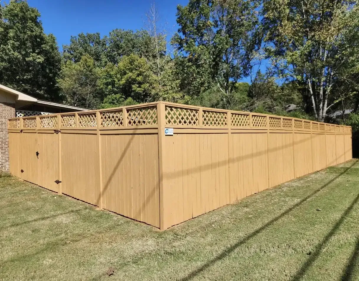 Tall tan wooden privacy fence enclosing a yard, with decorative lattice top, under a blue sky.