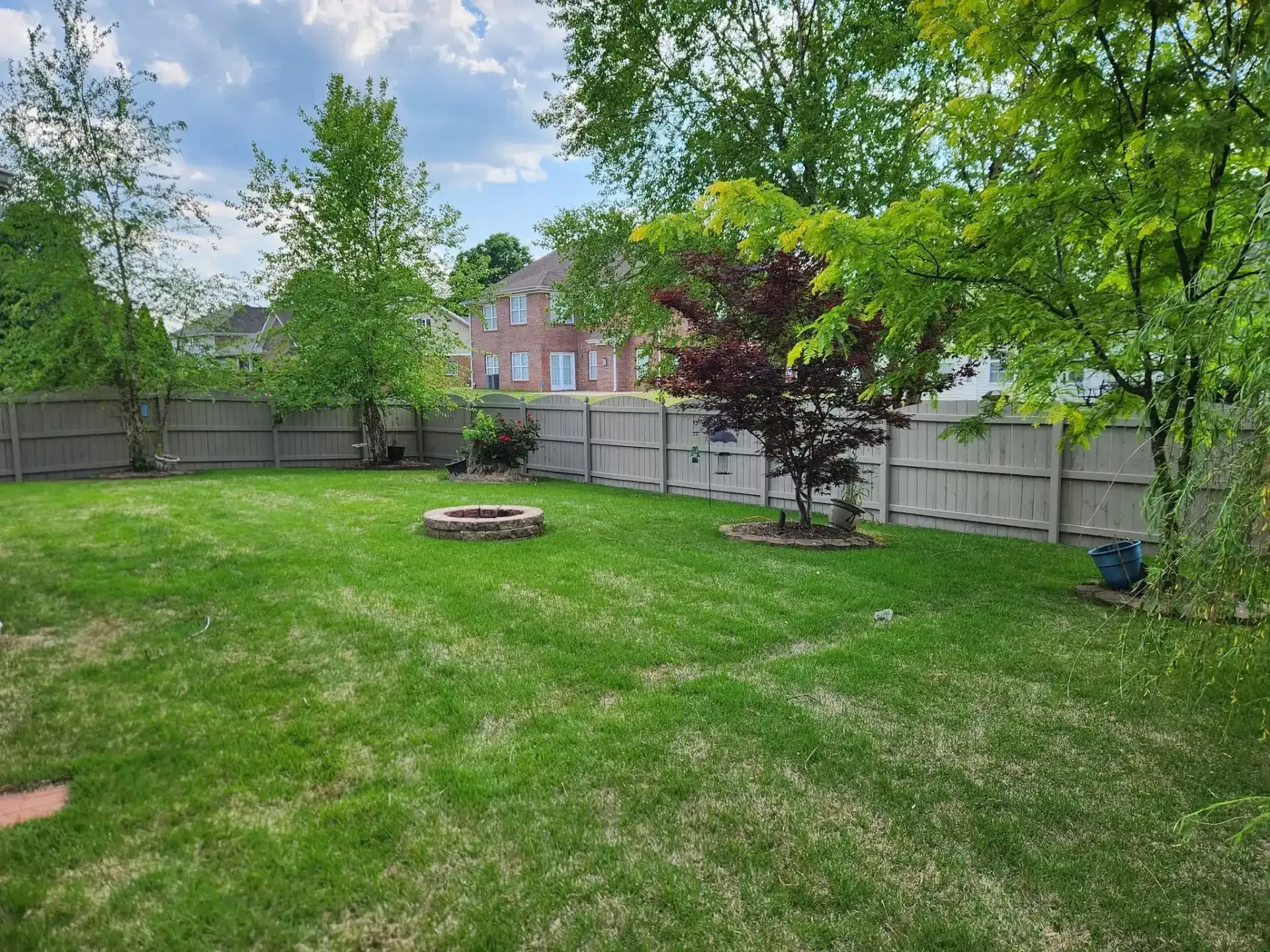 Backyard with green lawn, fire pit, trees, and a fenced perimeter under a partly cloudy sky