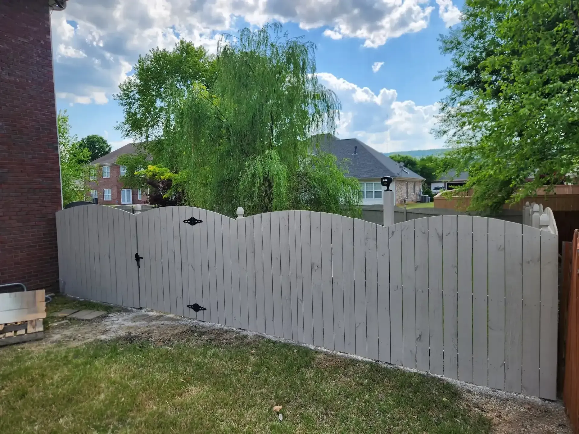 Gray wooden fence in a backyard with a grassy lawn, trees, and houses under a partly cloudy sky