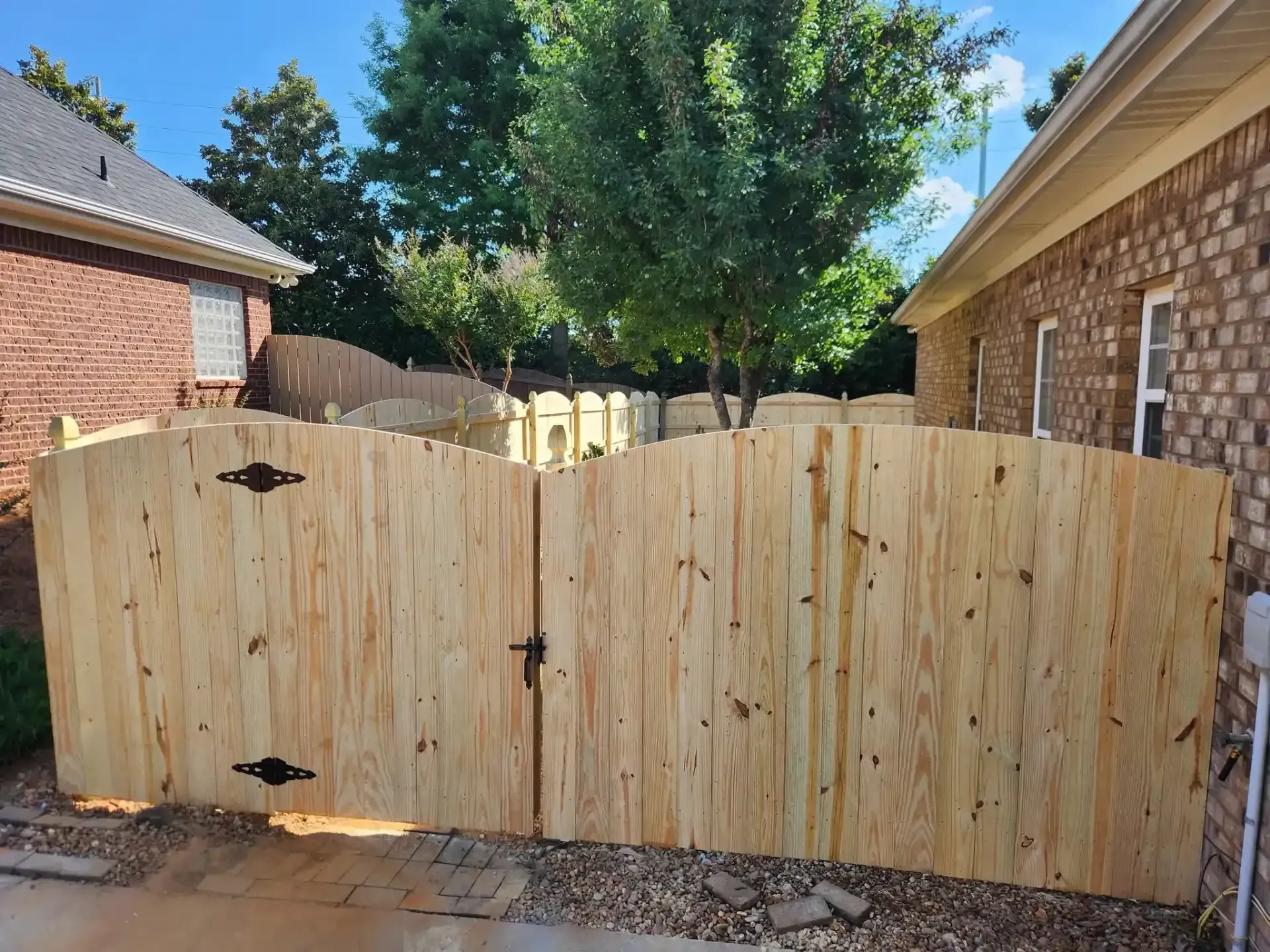 Wooden backyard gate between brick houses under bright sunlight, with a tree behind it.