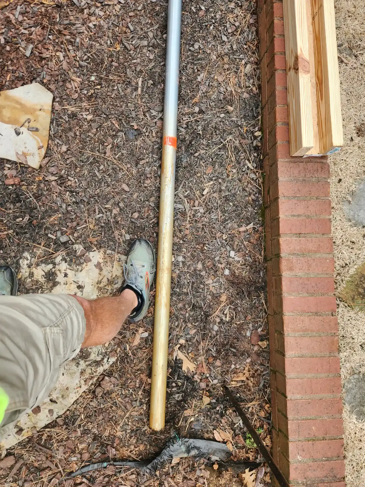 Outdoor gravel area with a person’s foot, a long metal pipe, and stacked wood beside a brick edge.