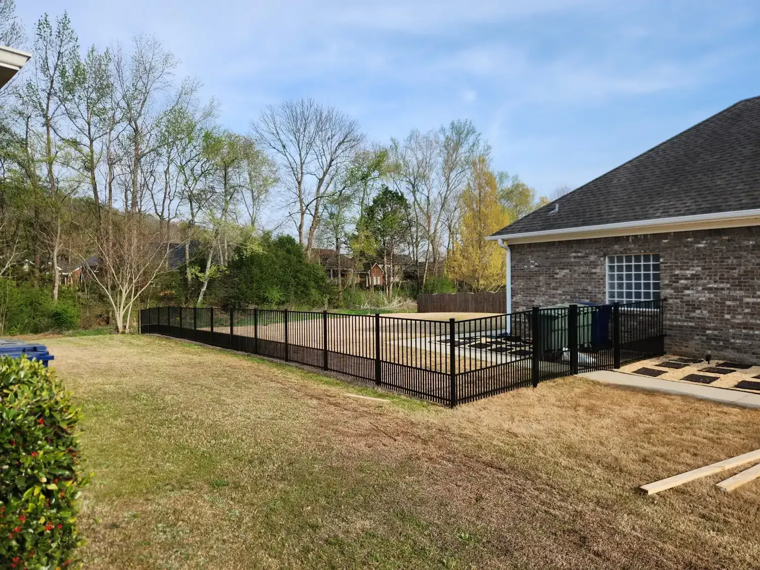 Backyard with a black metal fence and brick house patio beside a grassy lawn and trees