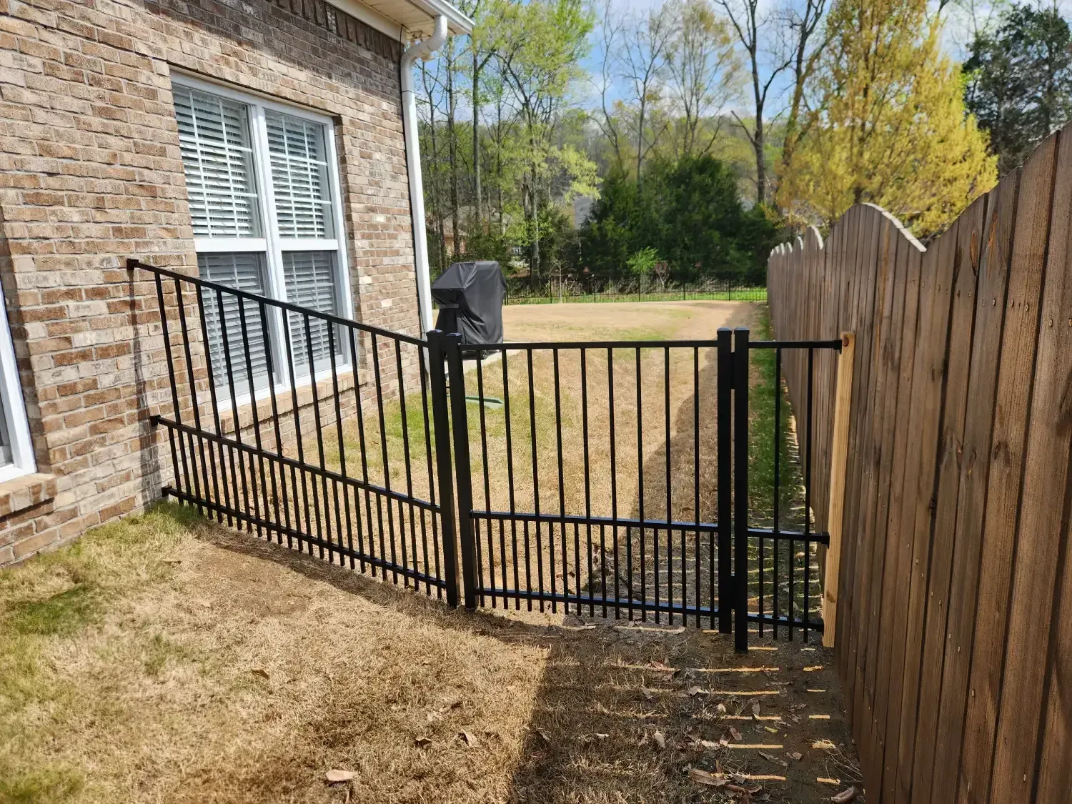 Black metal gate along a brick house and wooden fence beside a grassy yard.