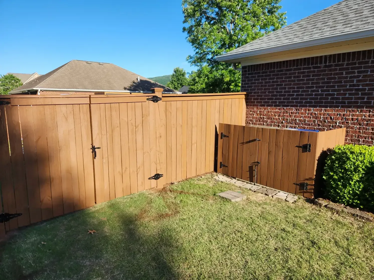 Backyard corner with wooden privacy fence, brick house wall, and a small trash bin by the grass