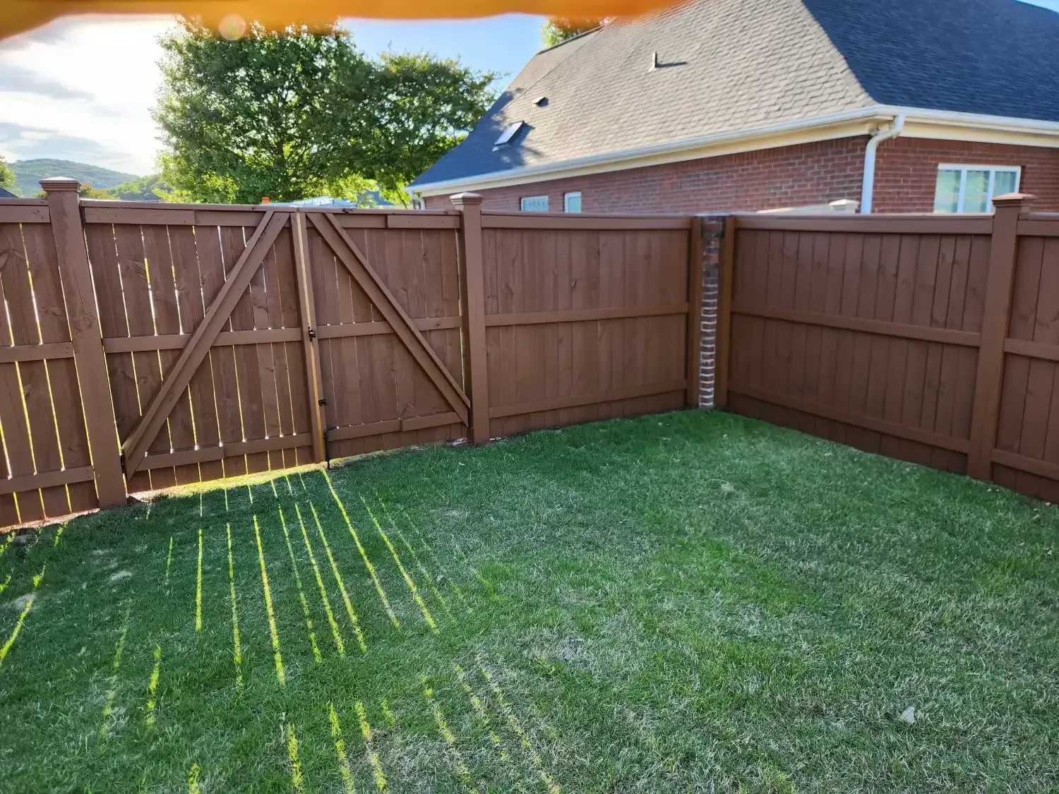 Small grassy backyard with a brown wooden fence and gate under a sunny sky.