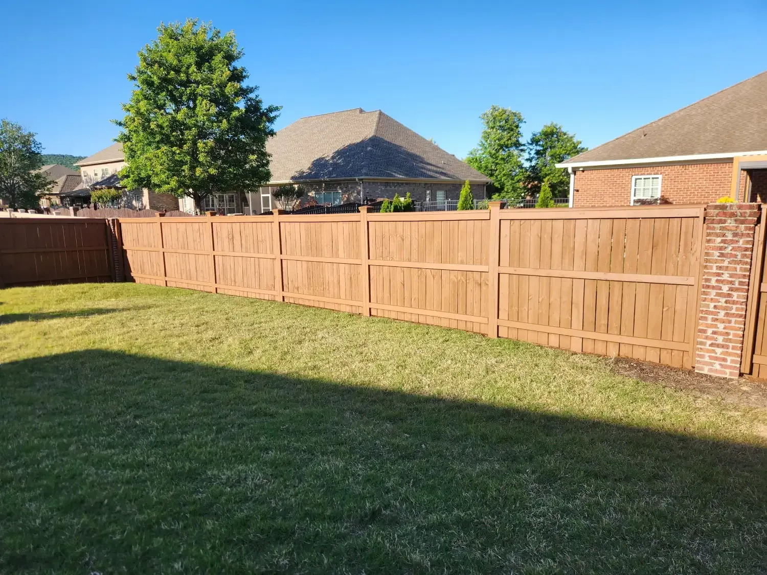 Sunny suburban backyard with green lawn, wooden privacy fence, brick pillars, and houses beyond