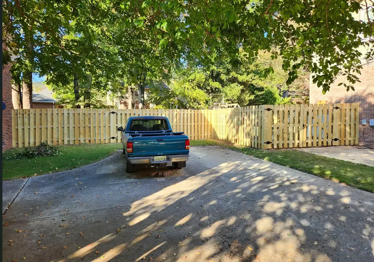Blue pickup truck parked on a sunlit driveway in front of a wooden fence and trees