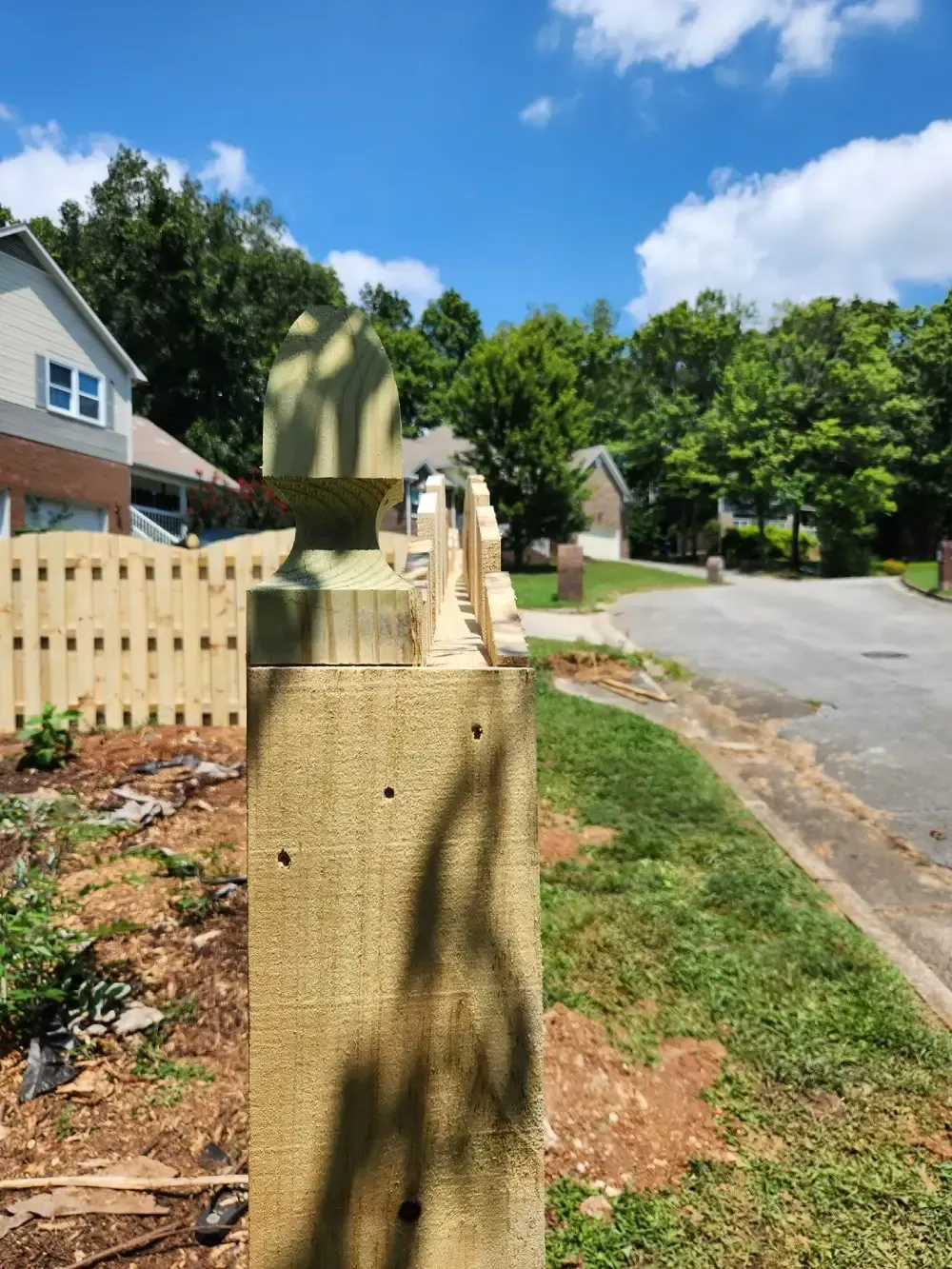 Residential street with a tan stone post, white fence, and trees under a blue sky
