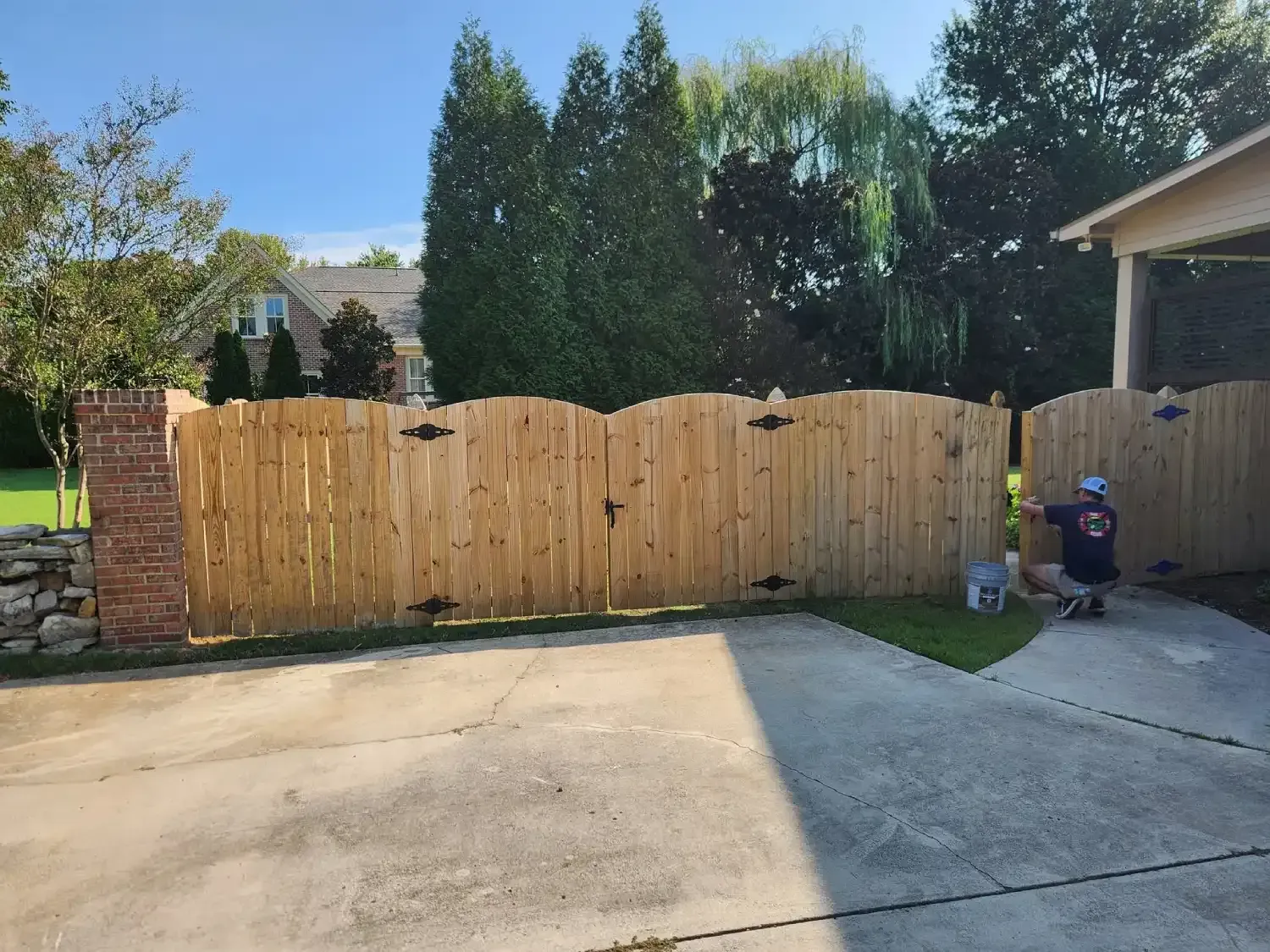 Wooden driveway gate beside a concrete driveway, with a small seated figure near the garage.