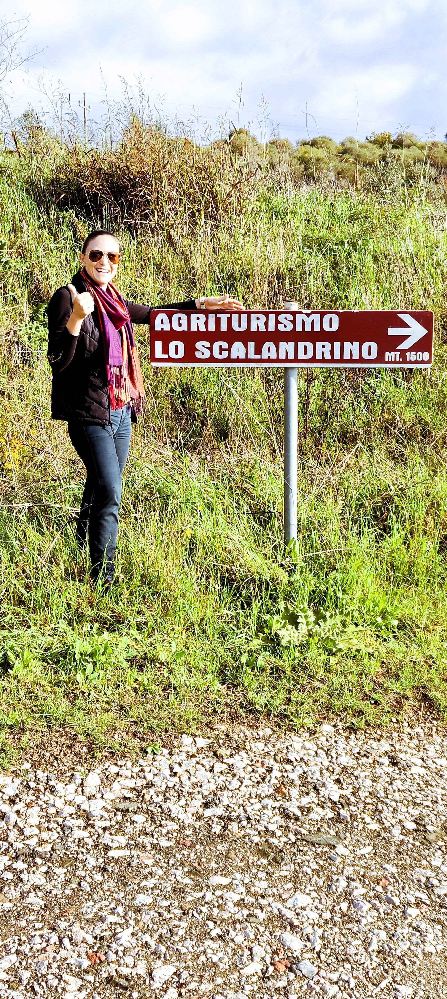 Ljuana stands beside a wooden post in a grassy meadow, looking at a red-and-white directional sign that reads “AGRITURISMO LO SCALANDRINO → MT. 1500,” with tall grass underfoot and trees in the background.
