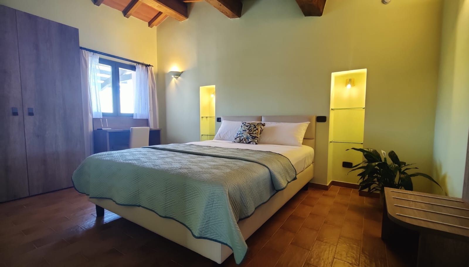 Bedroom with sloped wooden ceiling, king bed with quilt and white pillows between lit shelves, wardrobe by curtained window, tiled floor.