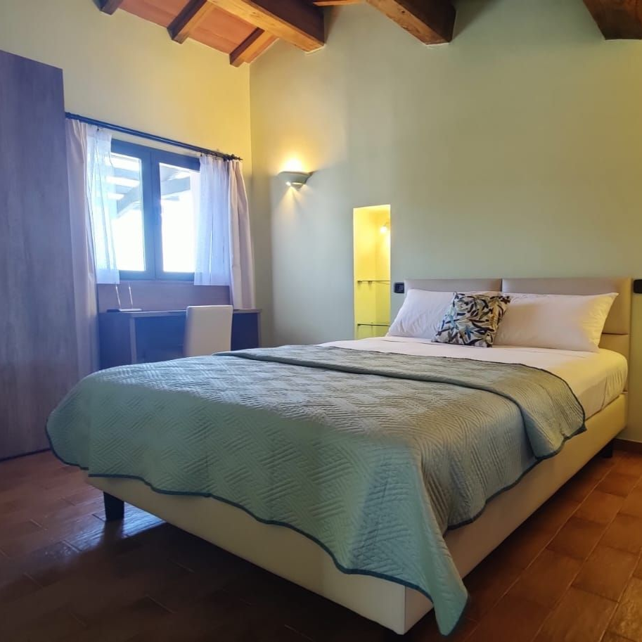Bedroom with sloped wooden ceiling, king bed with quilt and white pillows between lit shelves, wardrobe by curtained window, tiled floor.