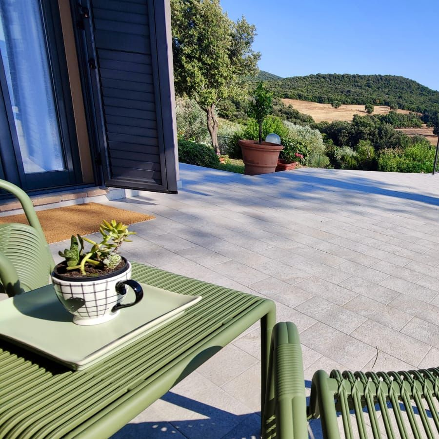Two light-green chairs flank a small white table holding a potted succulent on a pale tiled patio bordered by a black metal railing, overlooking rolling green hills, trees, and fields beneath a clear blue sky.