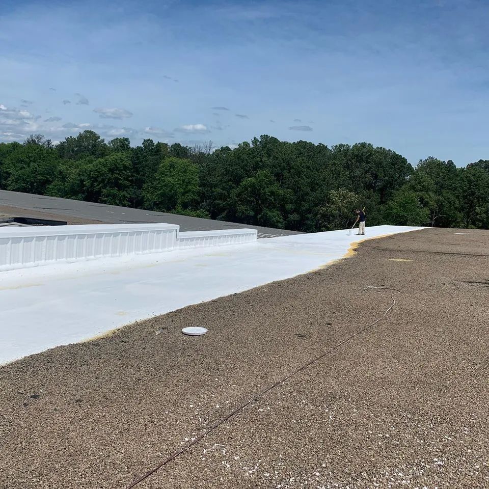 A roof with a new white coating next to a gravel area, trees in the background under a blue sky.