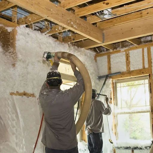 Workers installing insulation in a house, using a hose to spray the white material into walls and ceiling.