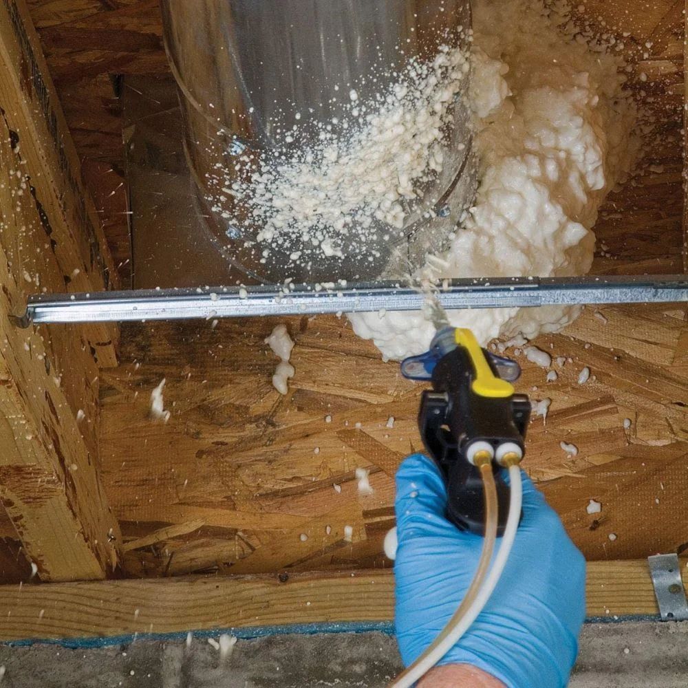 Person spraying expanding foam sealant around ductwork in a wood-framed space.
