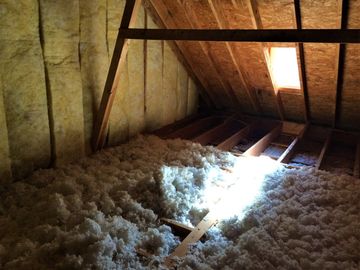 Attic with loose, white insulation covering the floor; wood beams and walls; sunlight streams in from a small window.