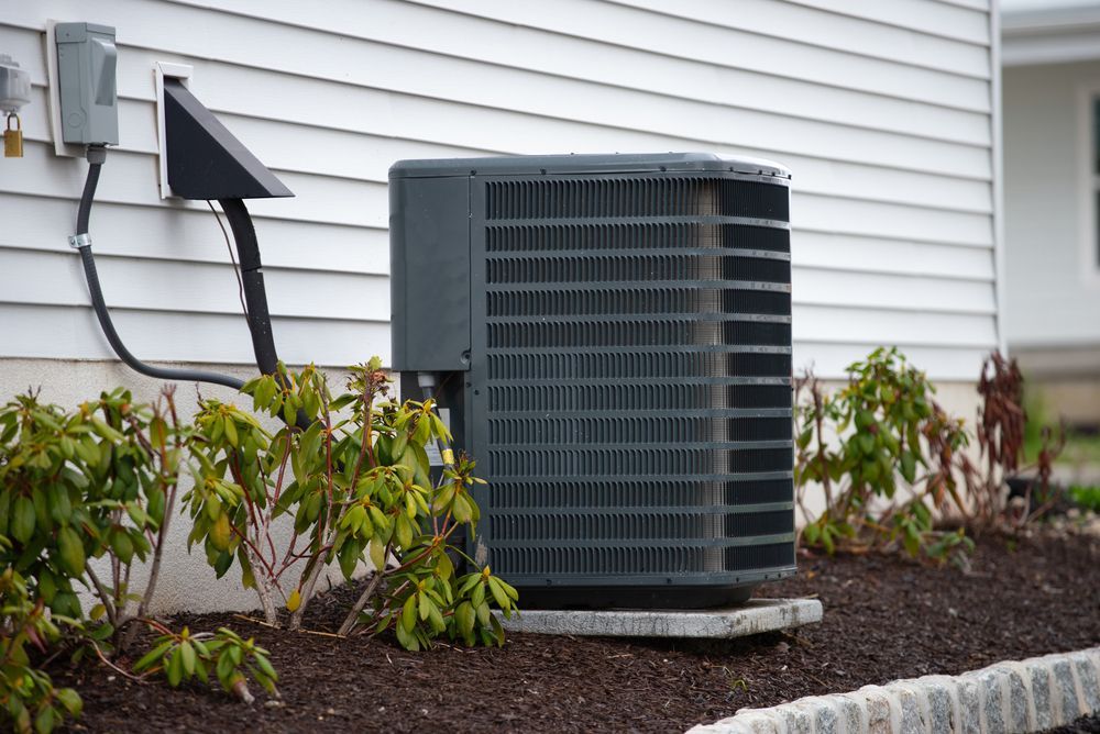 Gray air conditioning unit outside a white house next to small green bushes.