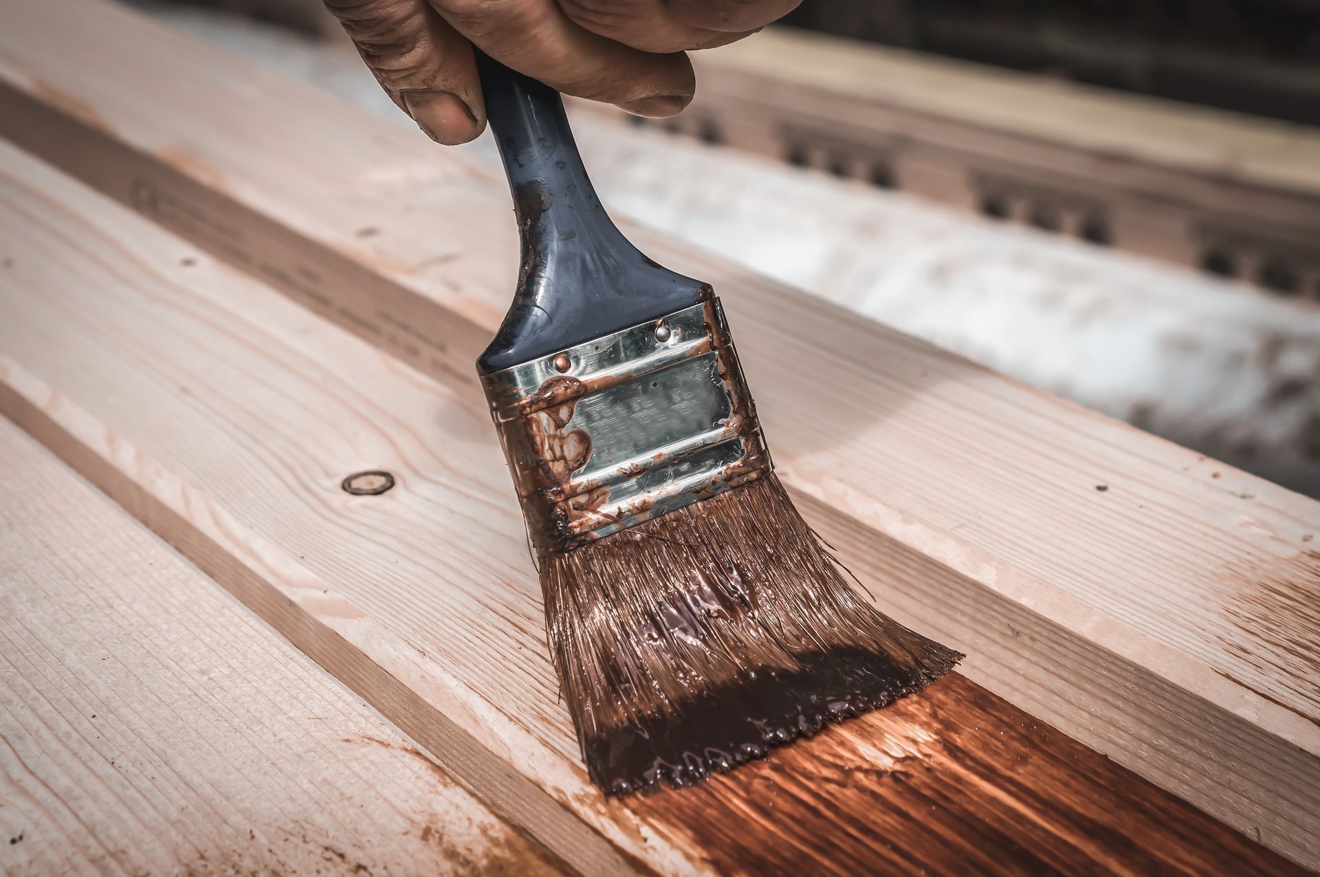 Hand applying brown stain to wooden planks with a paintbrush.