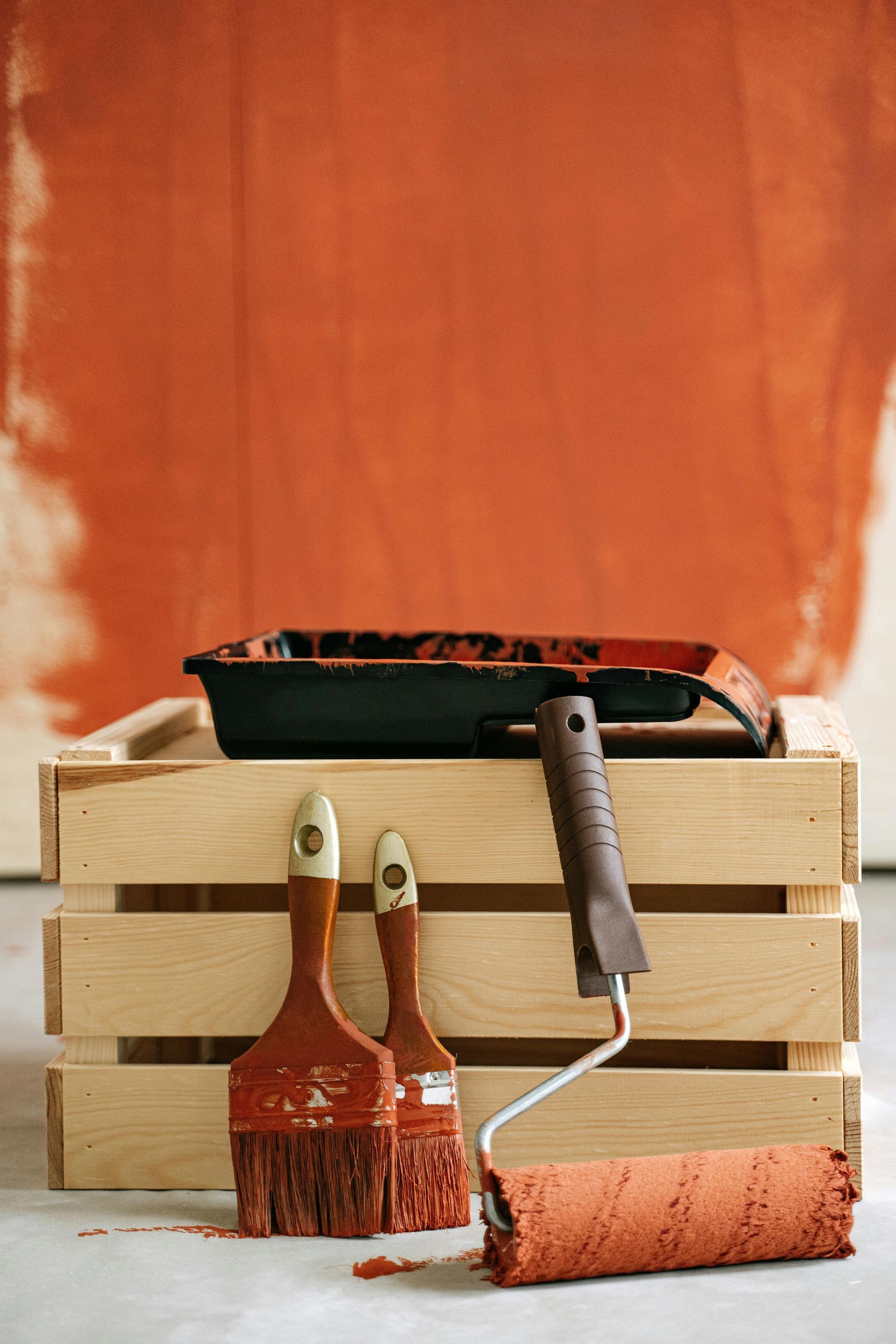 Paint supplies: paintbrushes, paint roller, and tray on a wooden crate, in front of a partially painted orange wall.