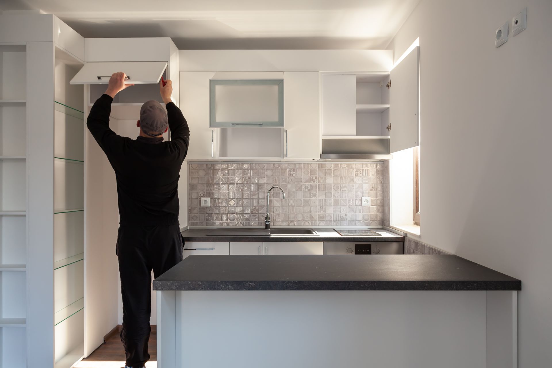 Person reaching up to install a white cabinet in a bright kitchen